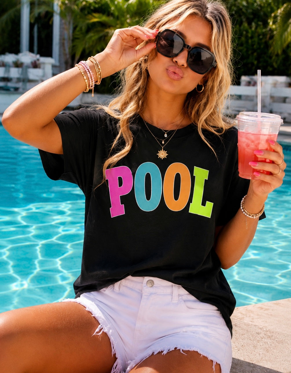 Woman in black 'POOL' t-shirt by a pool holding a colorful drink.