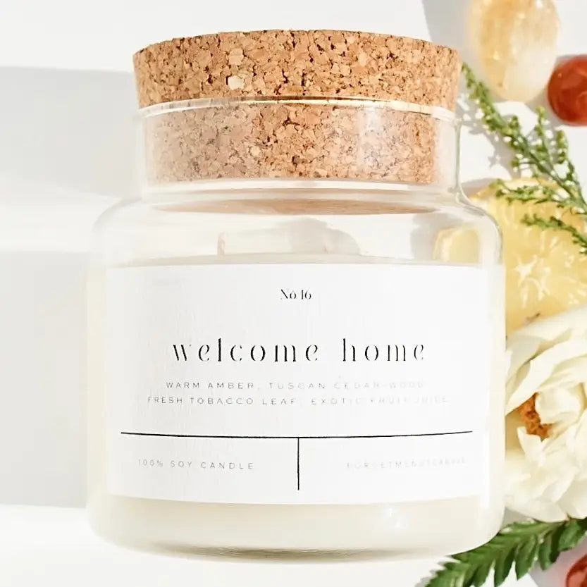 Candle jar with cork lid labeled 'welcome home' on a white background with flowers.