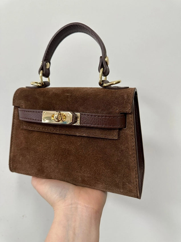 Brown suede handbag with gold hardware held by a hand against a gray background