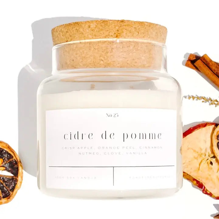 A clear glass jar candle with a cork lid and a white label reading 'cidre de pomme' and 'No 25', styled on a white background with dried citrus slices, cinnamon sticks, and a sprig of botanicals around it.