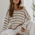 A woman wearing a beige and off-white horizontally striped knit sweater and white pants, seated indoors with a plant visible in the background.