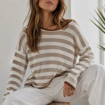 A woman wearing a beige and off-white horizontally striped knit sweater and white pants, seated indoors with a plant visible in the background.