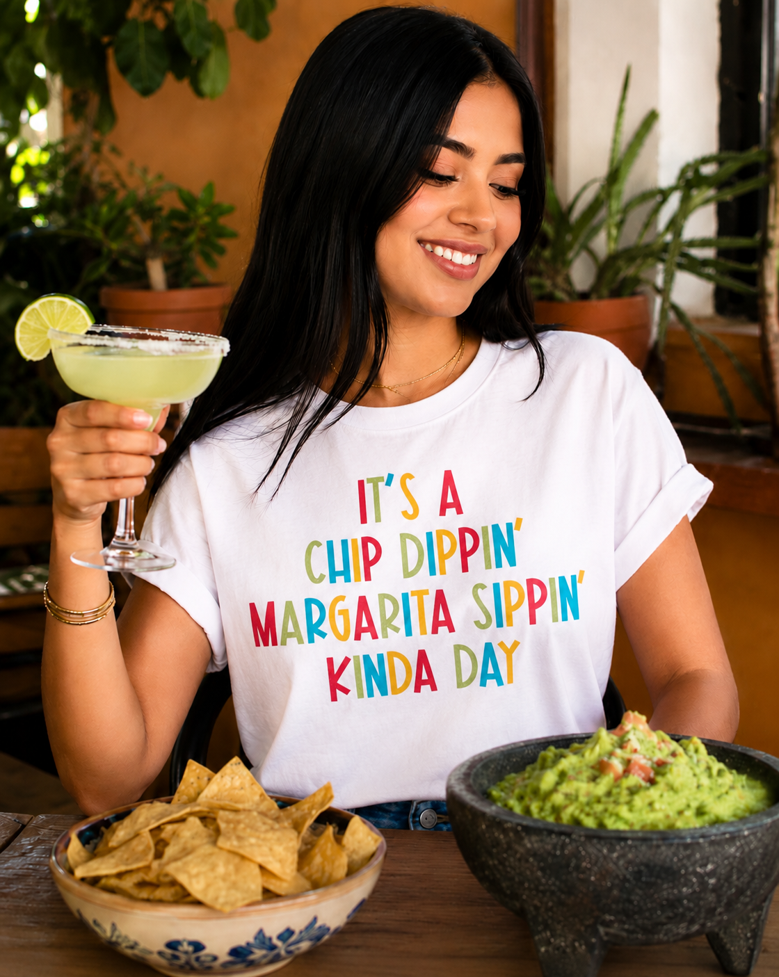 Woman in a white t-shirt with colorful text holding a margarita, surrounded by chips and guacamole.
