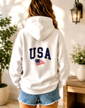 Person wearing a white hoodie with 'USA' and an American flag design, standing in a room with plants and a table.