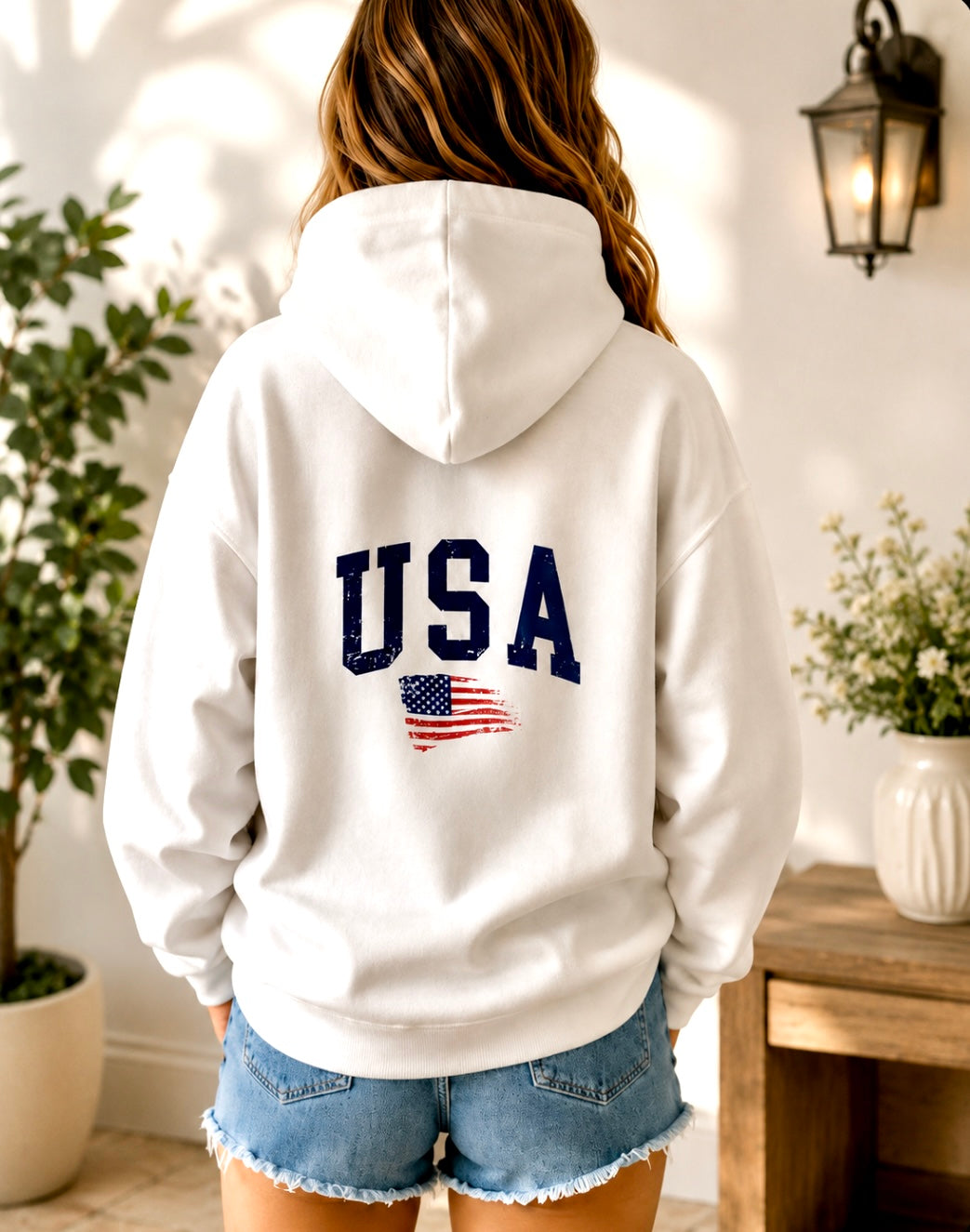 Person wearing a white hoodie with 'USA' and an American flag design, standing in a room with plants and a table.