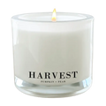 A lit white jar candle with a single flame, featuring a clear glass container and a black printed label that reads 'HARVEST' and 'PUMPKIN + PEAR' on a plain white background.
