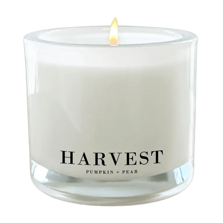 A lit white jar candle with a single flame, featuring a clear glass container and a black printed label that reads 'HARVEST' and 'PUMPKIN + PEAR' on a plain white background.