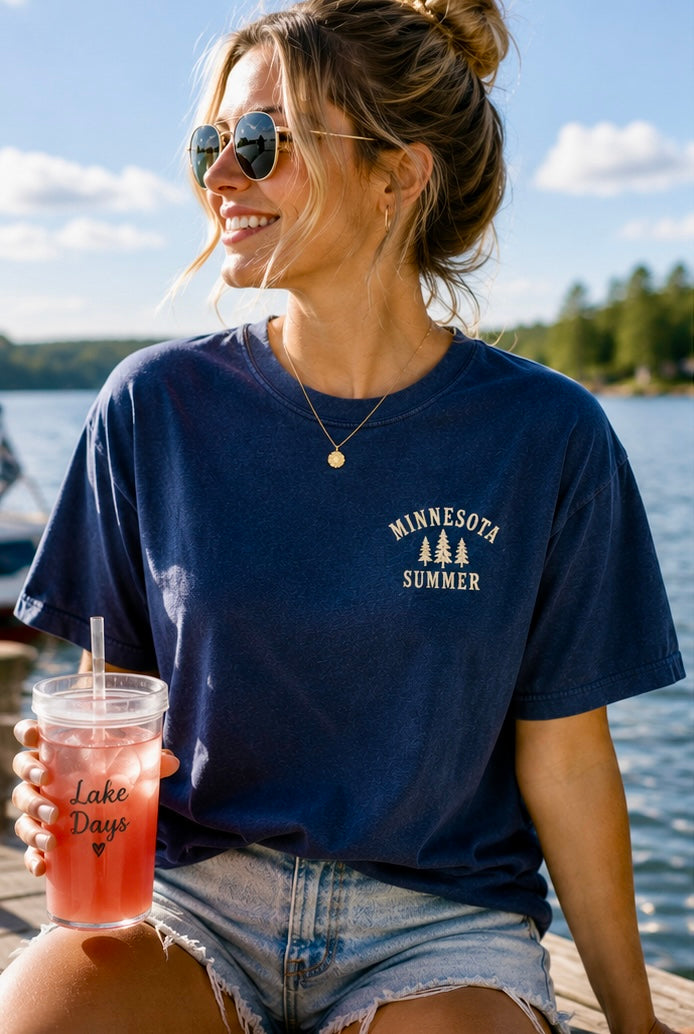 Woman wearing a navy blue 'Minnesota Summer' t-shirt by a lake.