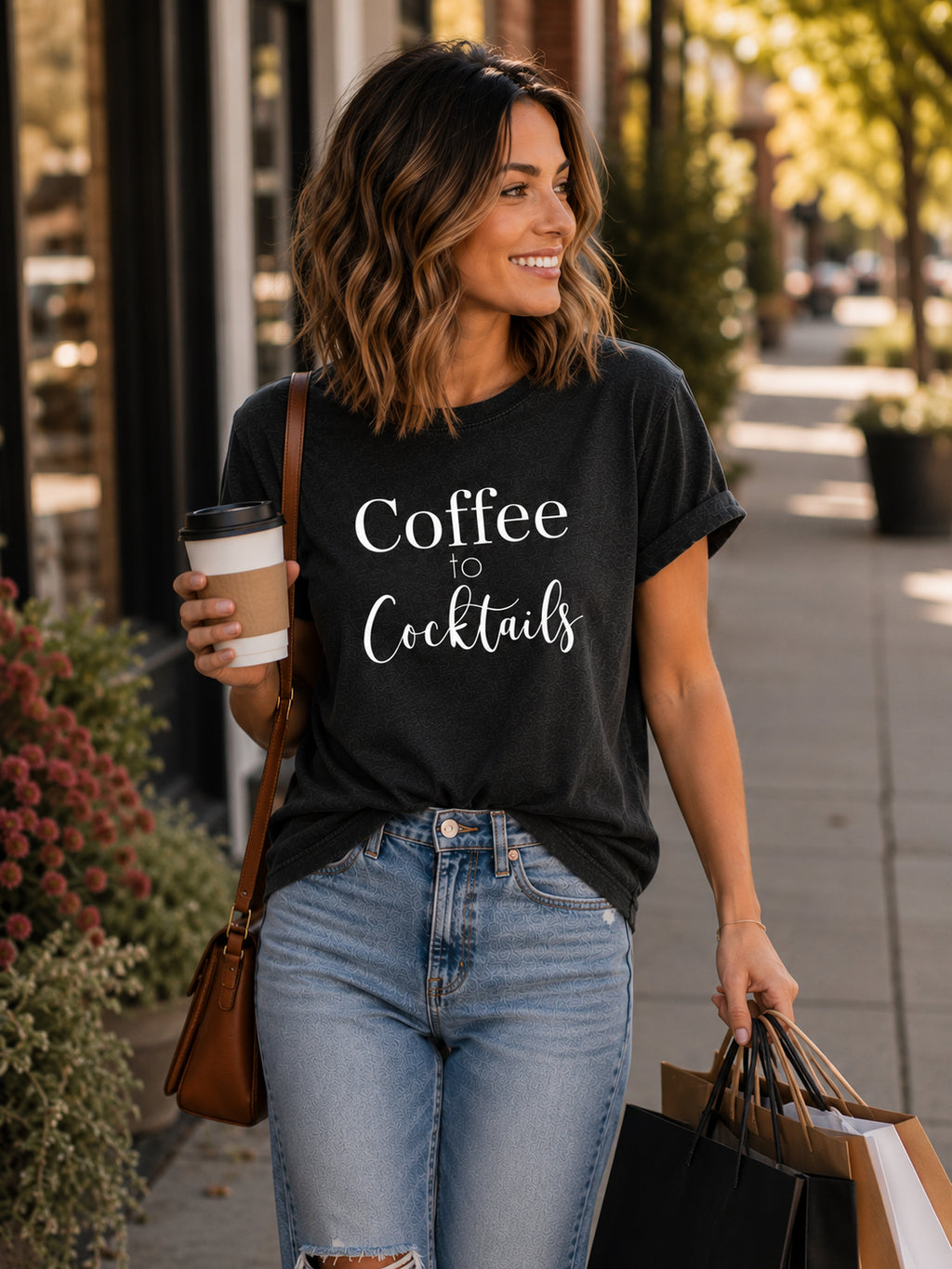 A woman walking on a city sidewalk wearing a black graphic t-shirt with the white text 'Coffee to Cocktails'. She is holding a takeaway coffee cup and shopping bags, with a brown shoulder bag slung over her shoulder.