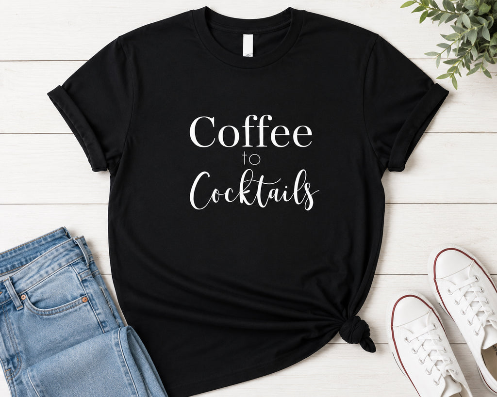 A black short-sleeve graphic T-shirt laid flat on a white wood background, styled with folded blue jeans, white sneakers, and a small green plant. The shirt has white text reading 'Coffee to Cocktails' across the front.