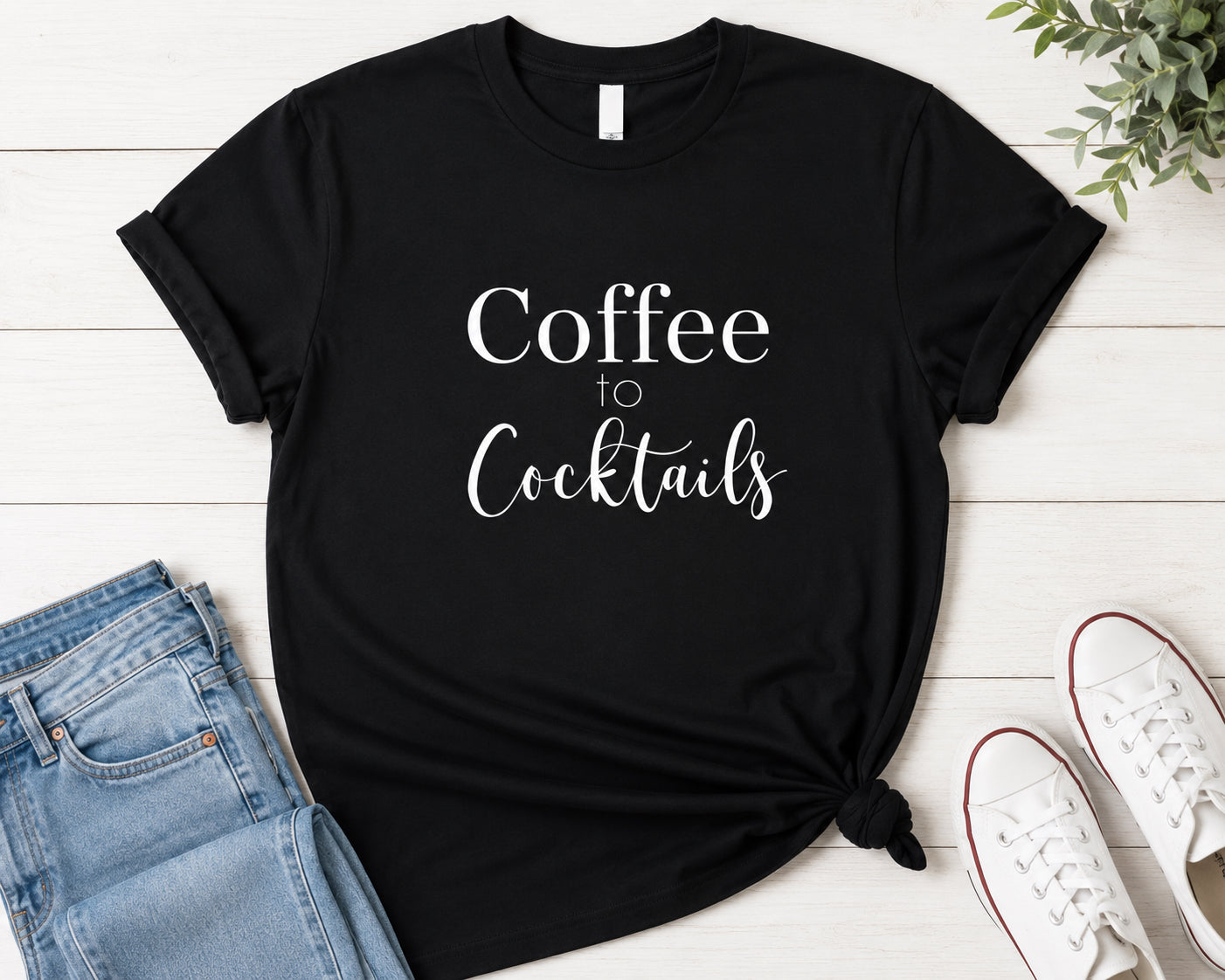 A black short-sleeve graphic T-shirt laid flat on a white wood background, styled with folded blue jeans, white sneakers, and a small green plant. The shirt has white text reading 'Coffee to Cocktails' across the front.