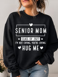 A woman wearing a black crewneck sweatshirt with a white graphic that reads 'SENIOR MOM CLASS OF 2027 I'M NOT CRYING, YOU'RE CRYING HUG ME' with small heart icons, paired with light blue jeans.