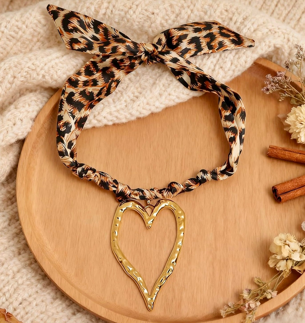 A leopard print fabric headband tied in a bow, featuring a gold heart-shaped pendant with rhinestone trim, displayed on a wooden tray with a cream knit blanket and dried flowers.