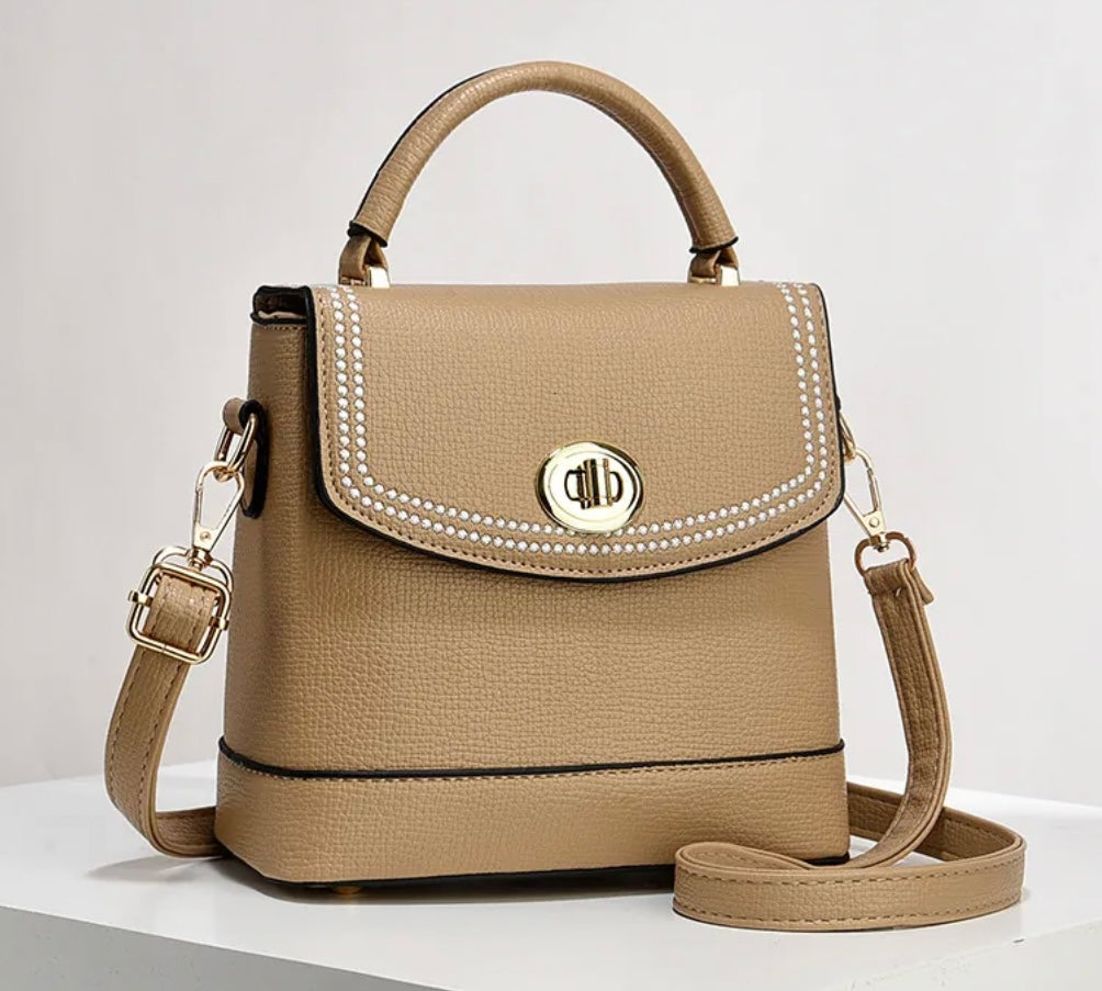 A structured beige faux-leather handbag with a top handle, a detachable shoulder strap, and a flap front decorated with white pearl-like studs and a round gold-tone logo clasp, shown on a white surface against a light gray background.