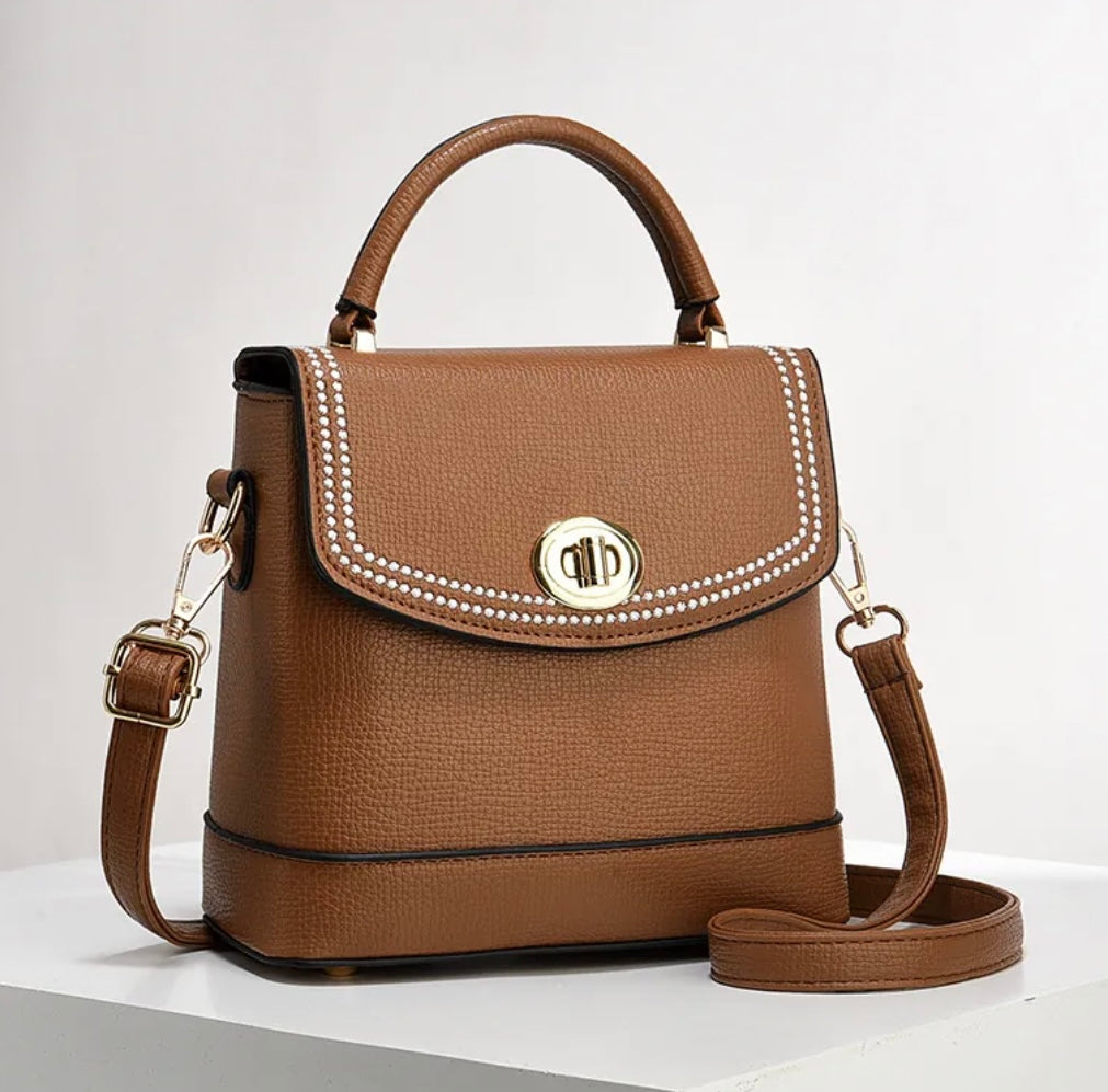 A brown pebbled leather handbag with a top handle, a detachable shoulder strap, and a silver-tone metal logo clasp on the front flap, displayed on a white surface against a light gray background.