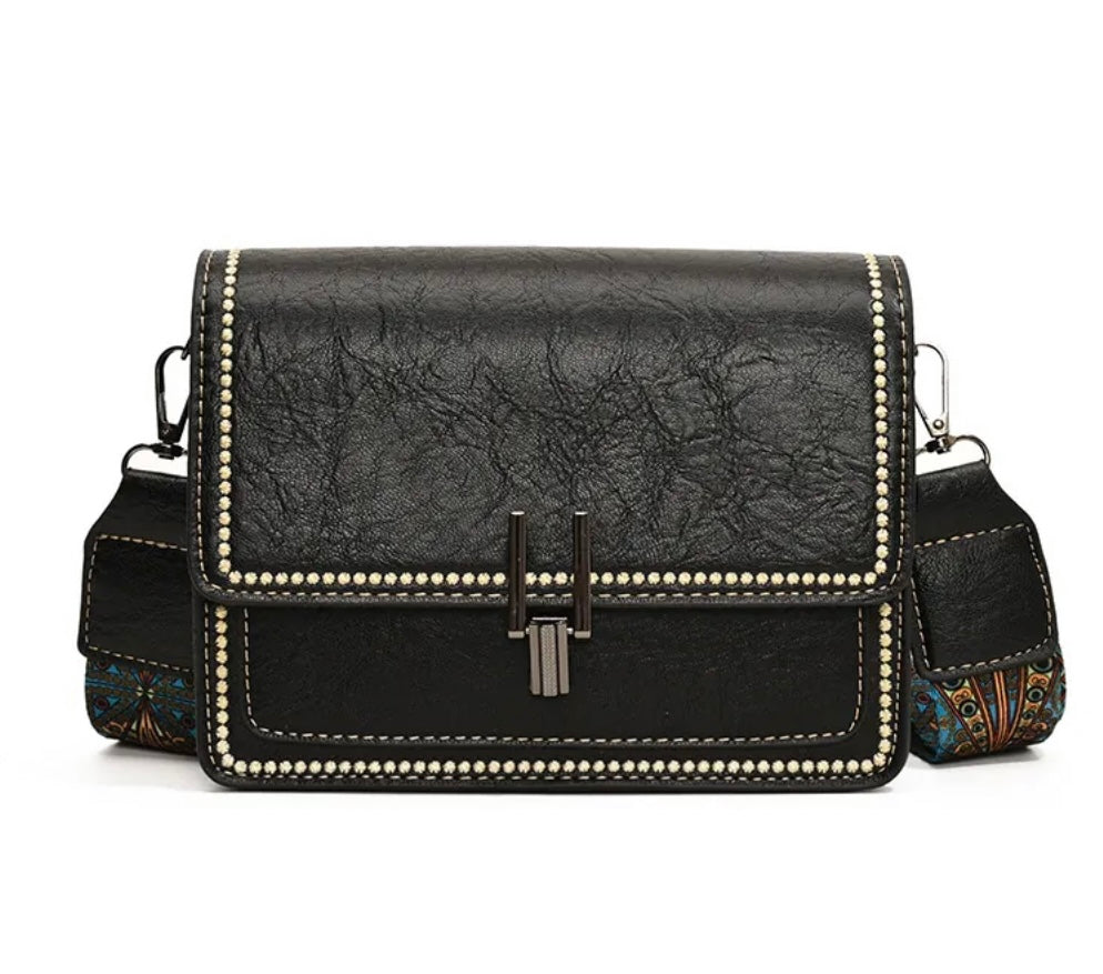 Black faux-leather shoulder bag with a textured finish, decorative silver-tone studs around the edges, a central metal clasp, and a patterned fabric side pocket, shown on a white background.