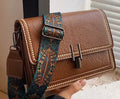 A brown faux-leather shoulder bag with a decorative blue and gold patterned strap, held by a hand in front of a window with blinds.