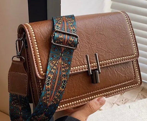 A brown faux-leather shoulder bag with a decorative blue and gold patterned strap, held by a hand in front of a window with blinds.