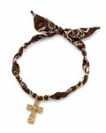 A brown silk scarf necklace with a white and brown pattern, tied in a knot at the top and featuring a dangling gold cross pendant.