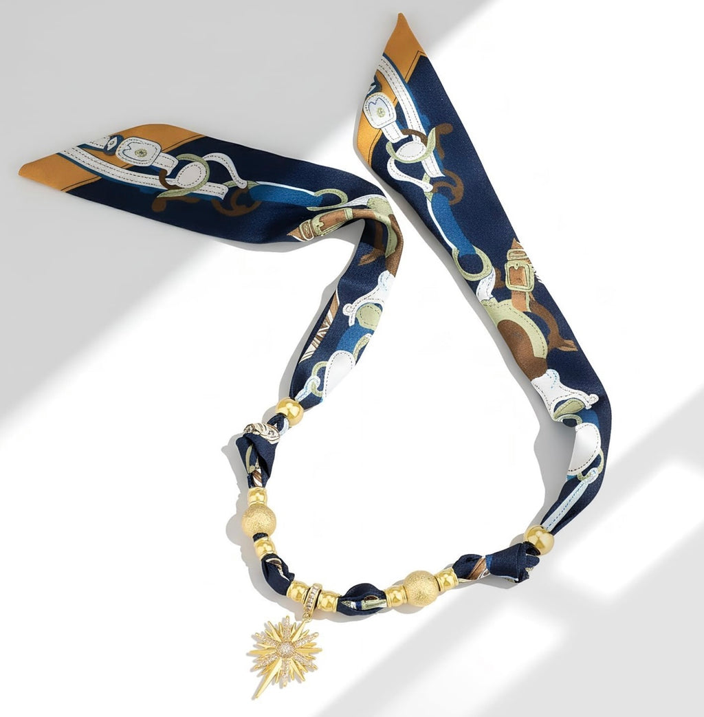 A luxury silk scarf styled as a necklace with gold-toned beads and a starburst charm, featuring a navy blue, gold, and white horse-and-carriage pattern.