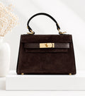 A dark brown suede top-handle handbag with gold-tone hardware, displayed on a white pedestal next to a white vase with dried flowers.