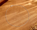 A delicate silver chain necklace with small spaced beads, arranged in a circle on a textured brown surface with soft shadows.