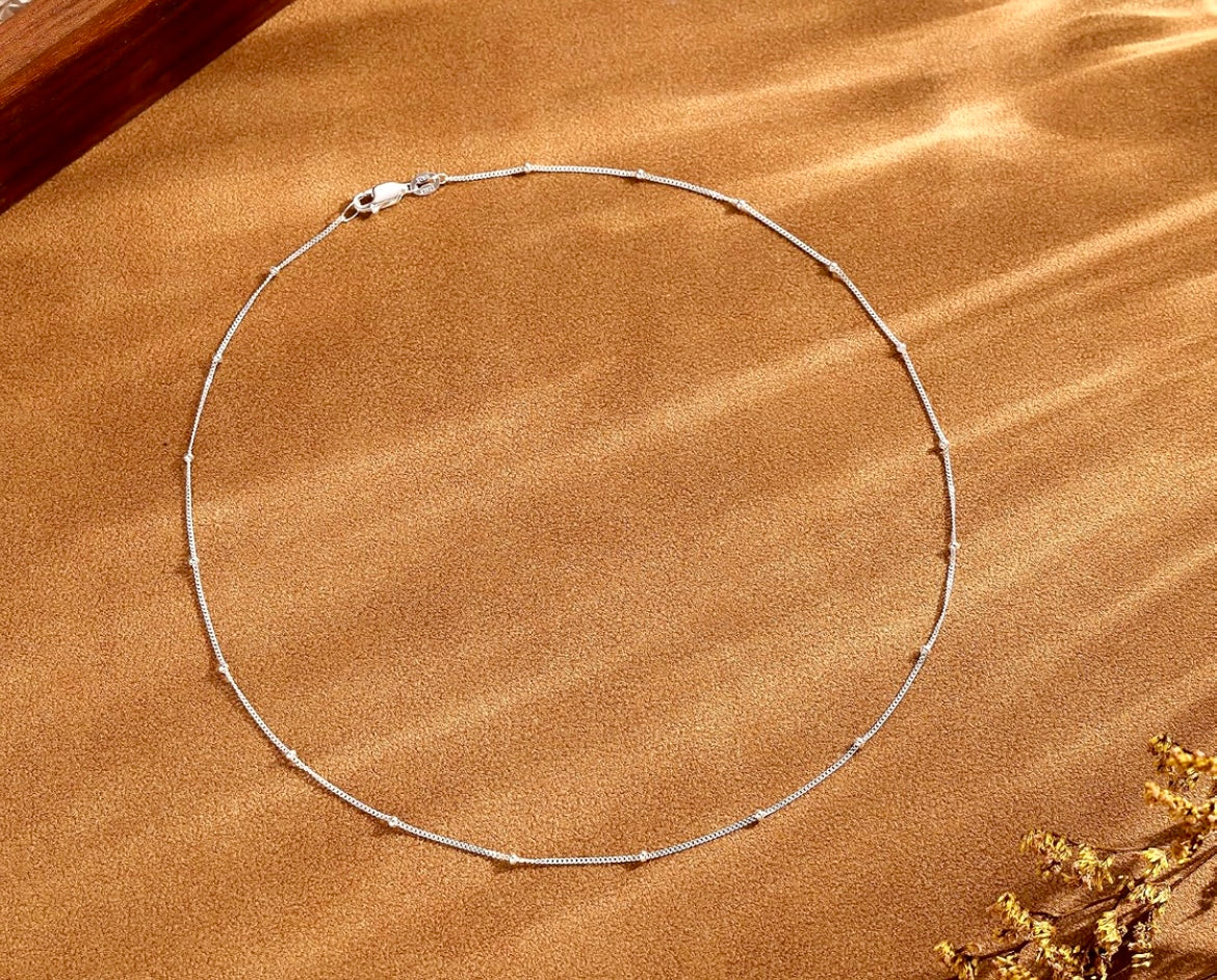 A delicate silver chain necklace with small spaced beads, arranged in a circle on a textured brown surface with soft shadows.