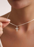 A close-up of a delicate silver chain necklace with a small cross pendant, worn on a model's neck with a hand gently holding the chain.