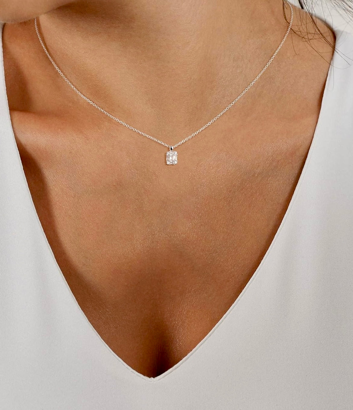 A close-up of a model wearing a delicate silver chain necklace with a small rectangular clear gemstone pendant, styled with a white V-neck top.