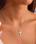 A close-up of a model wearing a delicate silver chain necklace with a small, minimalist cross pendant.
