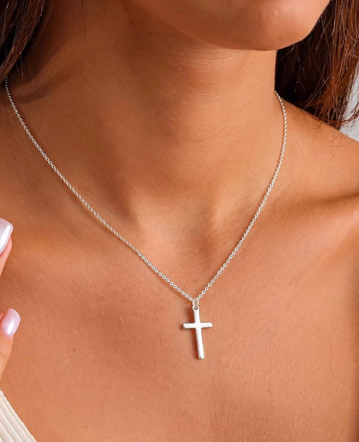 A close-up of a model wearing a delicate silver chain necklace with a small, minimalist cross pendant.