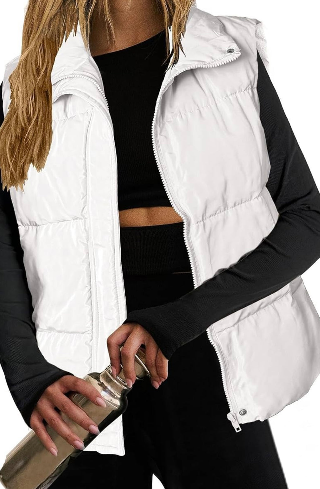 A close-up of a model wearing a white puffer vest over a black long-sleeve crop top and black pants, holding a metallic bottle against a plain white background.