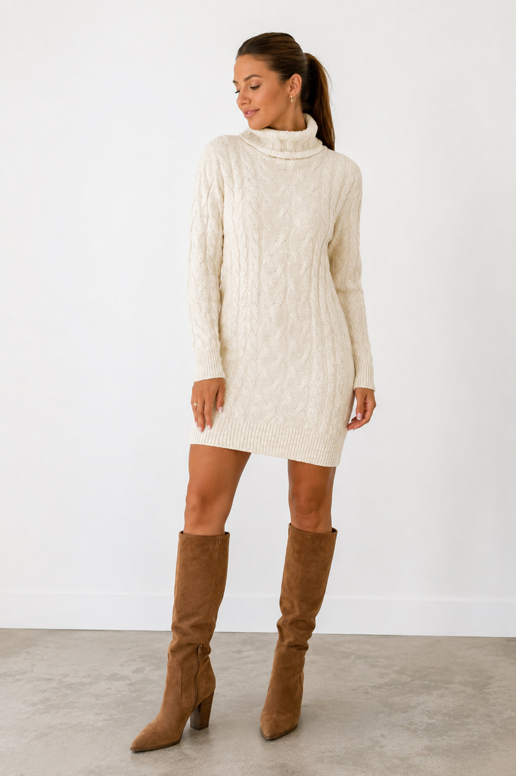 A woman modeling a cream cable-knit mock-neck mini sweater dress with brown suede knee-high boots, standing against a white studio wall.