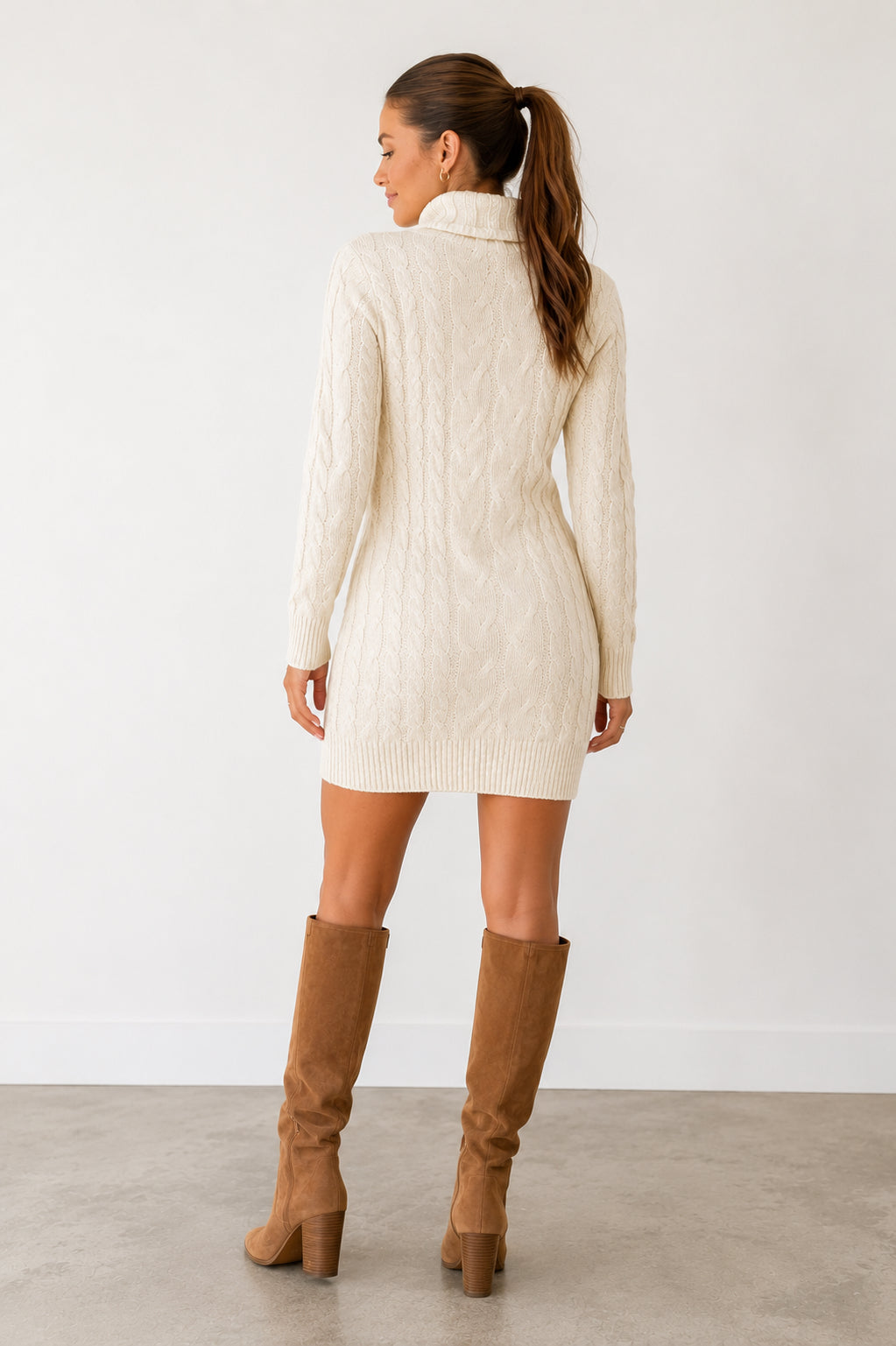 A woman shown from the back wearing a cream cable-knit mock-neck mini dress and brown knee-high boots, standing against a plain white wall.