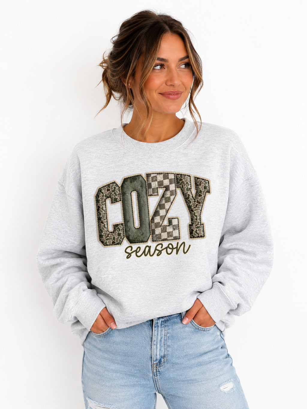 A woman wearing a light gray crewneck sweatshirt with a graphic that reads 'COZY season' in textured block letters, paired with light blue jeans, against a plain white background.