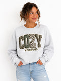 A woman wearing a light gray crewneck sweatshirt with a graphic that reads 'COZY season' in textured block letters, paired with light blue jeans, against a plain white background.