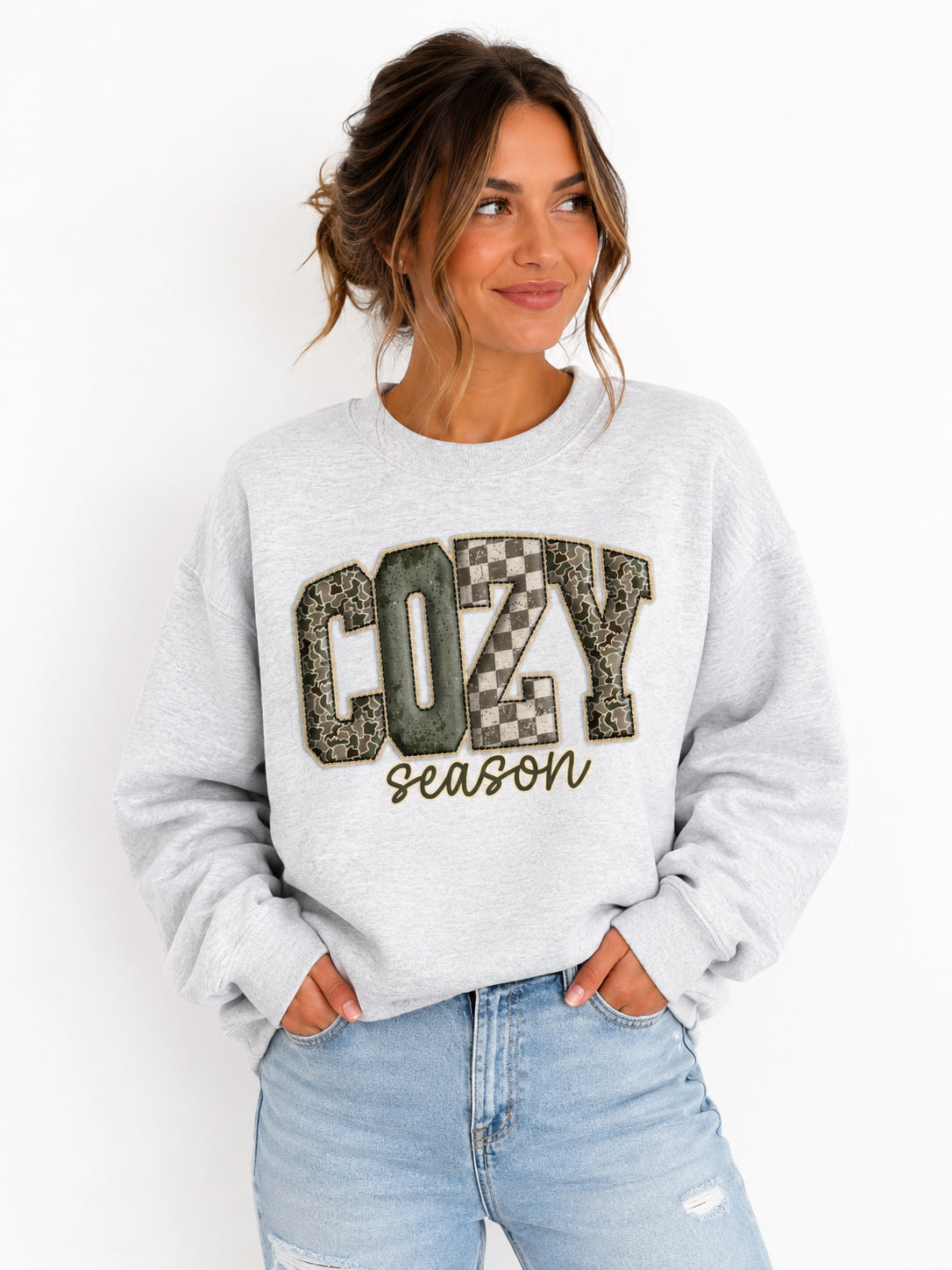 A woman wearing a light gray crewneck sweatshirt with a graphic that reads 'COZY season' in textured block letters, paired with light blue jeans, against a plain white background.