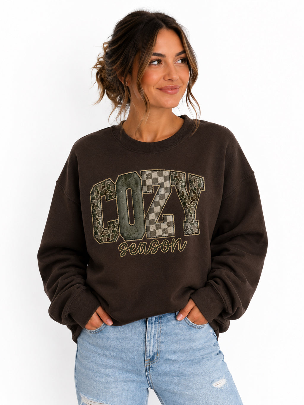 A woman wearing a dark brown crewneck sweatshirt with a graphic that reads 'COZY SEASON' in camouflage and checkered patterns, paired with light blue jeans, against a plain white background.