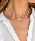 A close-up of a model wearing a delicate silver beaded necklace with a small cross charm, styled with a white collared shirt.