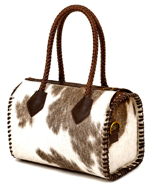A cowhide duffel bag with brown braided handles and braided trim, featuring a natural black and white spotted pattern on a white background.