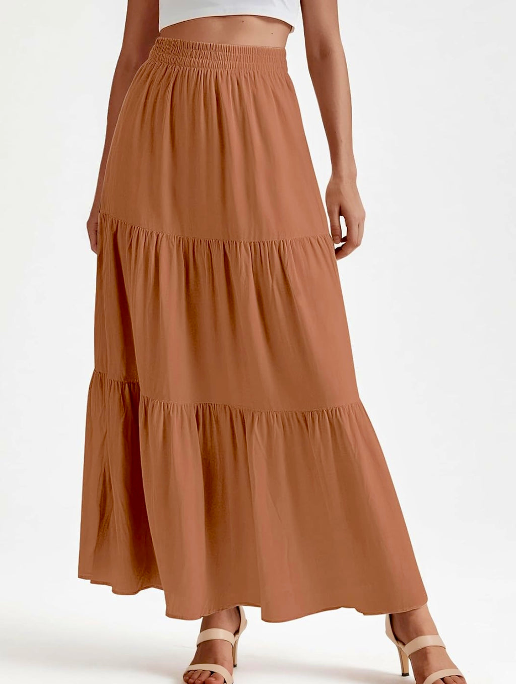 A woman wearing a tiered maxi skirt in a warm camel brown color, paired with a white crop top and beige strappy heels, shown against a plain white studio background.