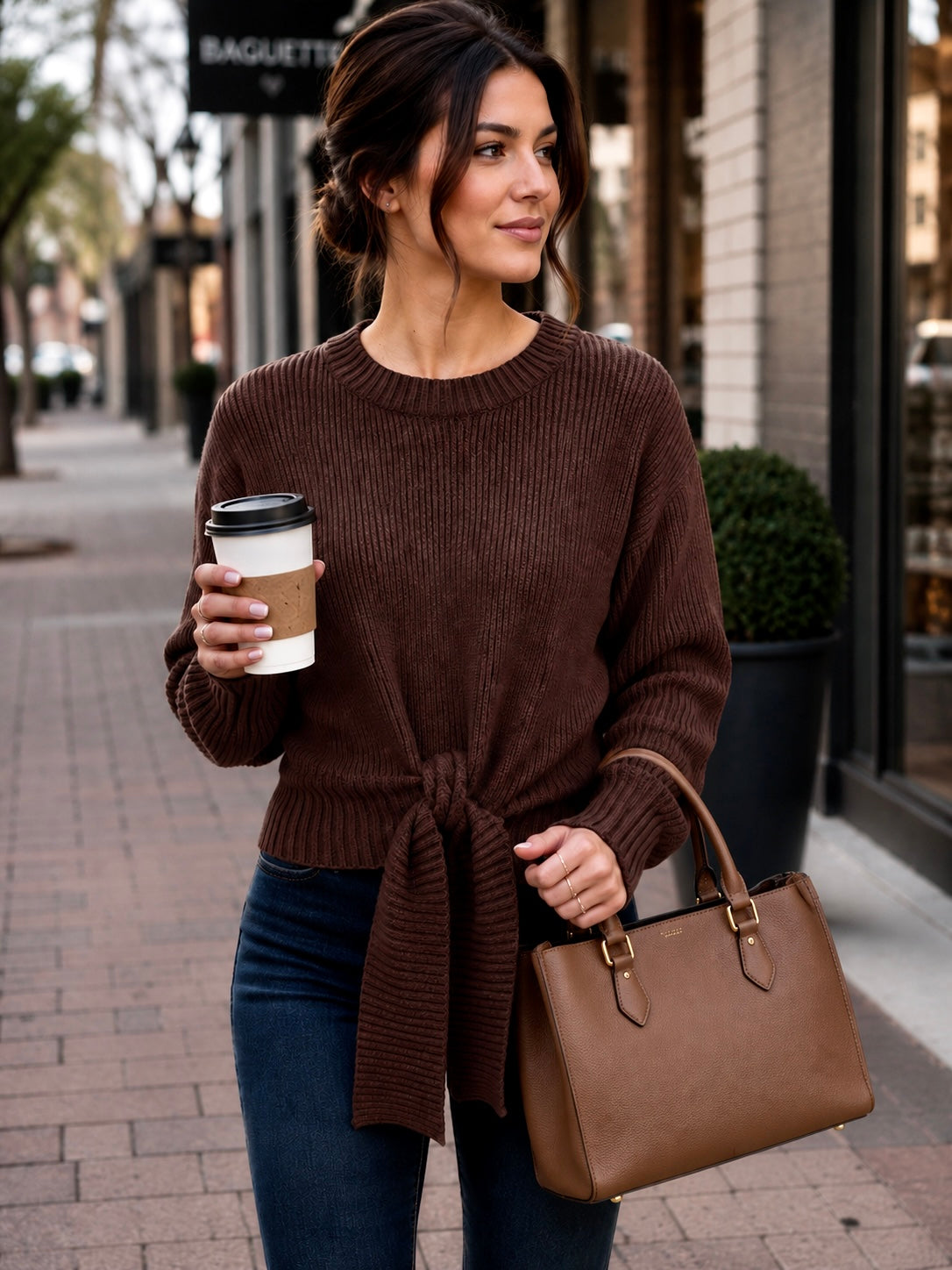 A woman stands on a city sidewalk wearing a brown ribbed knit sweater tied at the waist, dark blue jeans, and carrying a brown leather handbag while holding a takeaway coffee cup.