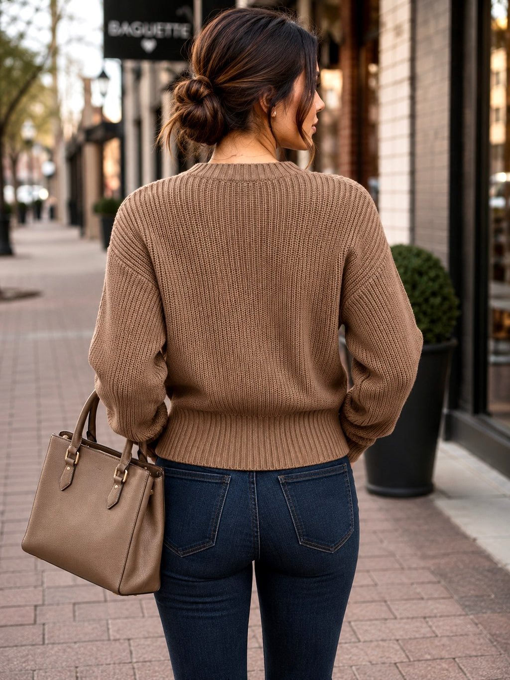A woman viewed from behind wearing a taupe brown ribbed knit sweater and dark blue jeans, carrying a taupe leather handbag on a city sidewalk.