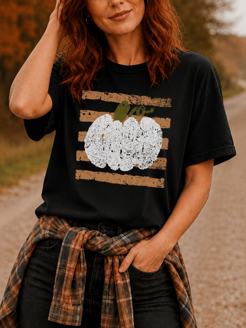 A black short-sleeve t-shirt with a distressed autumn graphic featuring horizontal brown stripes and a white pumpkin styled on a woman wearing jeans in a wooded background.