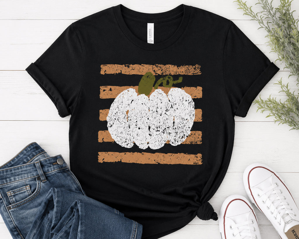 A black short-sleeve t-shirt with a distressed autumn graphic featuring horizontal brown stripes and a white pumpkin with a green stem, styled in a flat lay with blue jeans, white sneakers, and green foliage on a white wood background.