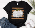 A black short-sleeve t-shirt with a distressed autumn graphic featuring horizontal brown stripes and a white pumpkin with a green stem, styled in a flat lay with blue jeans, white sneakers, and green foliage on a white wood background.