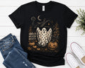 A black short-sleeve graphic T-shirt laid flat on a white wood surface, styled with blue jeans, white sneakers, and a small plant. The shirt features a Halloween-themed illustration with a ghost, pumpkins, a haunted house, a crescent moon, and the words 'ALL LIVE WHERE NOE'.