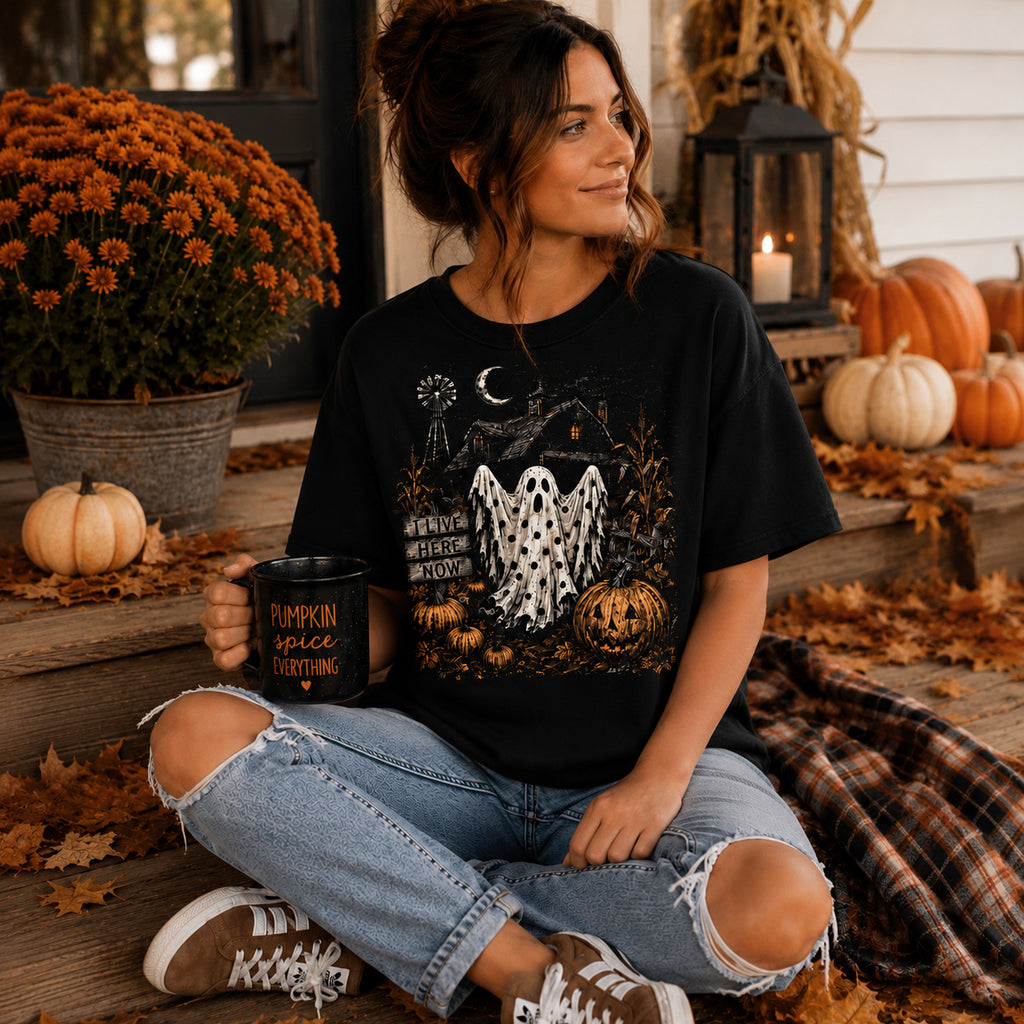 A woman sits cross-legged on a fall-themed porch wearing a black graphic t-shirt with a ghost and pumpkin design, paired with ripped blue jeans and brown sneakers. She holds a black mug with a 'Pumpkin Spice Everything' design.