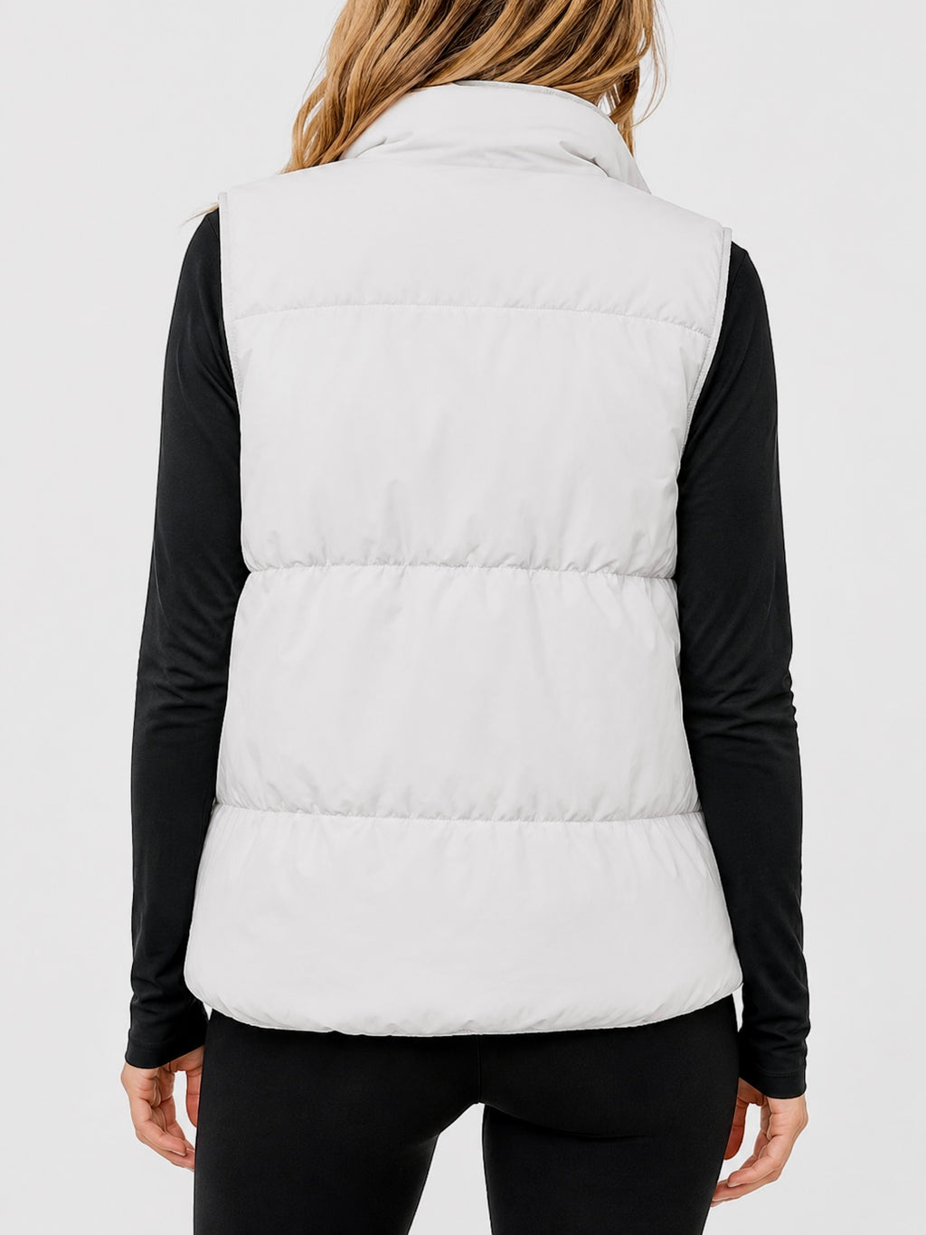 A woman shown from the back wearing a white puffer vest over a black long-sleeve top and black pants, against a light gray studio background.