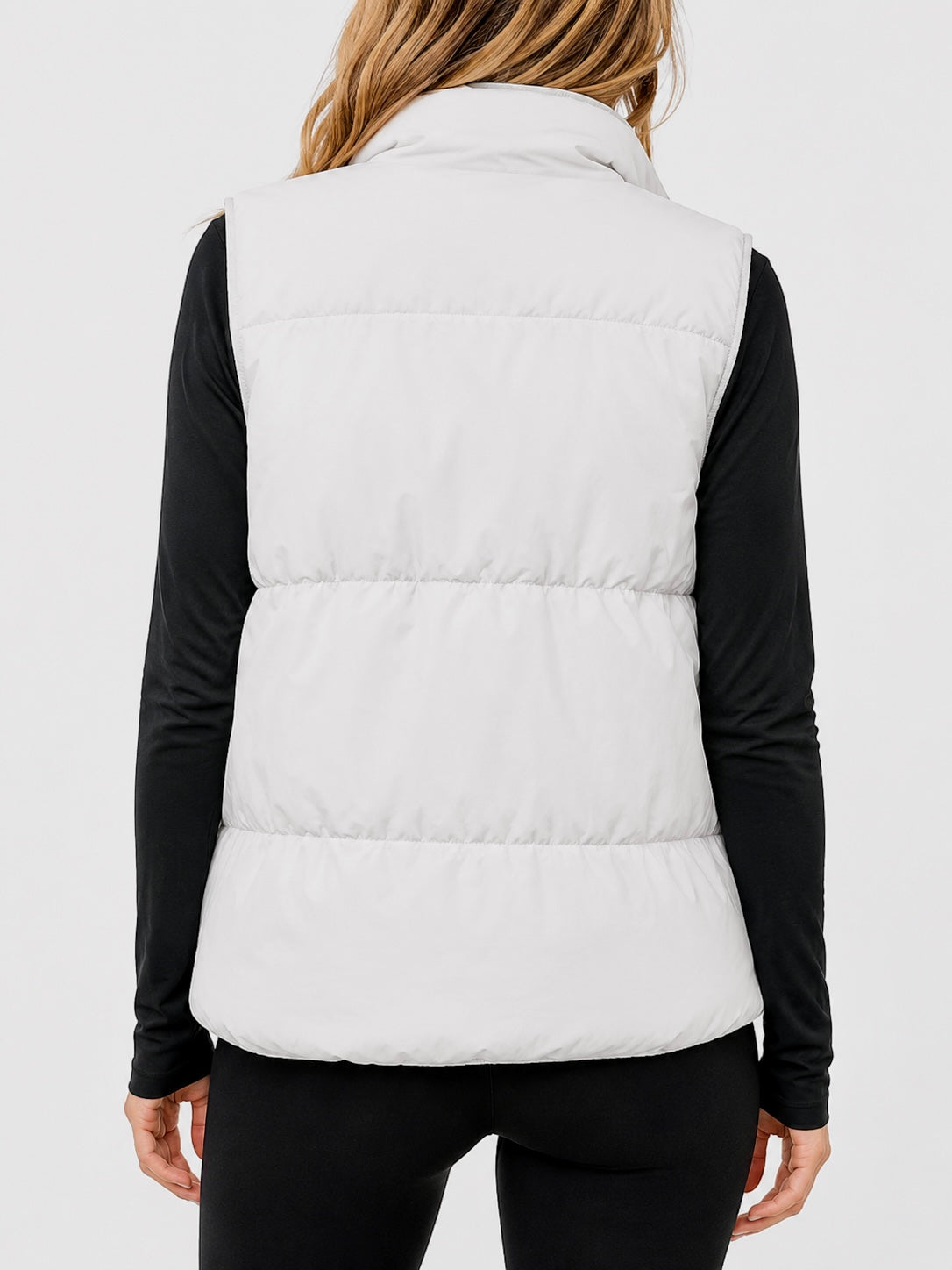 A woman shown from the back wearing a white puffer vest over a black long-sleeve top and black pants, against a light gray studio background.